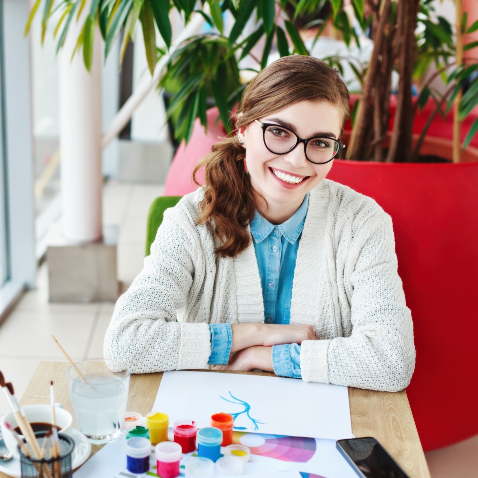 Cute girl wearing white sweater with glasses sitting in cafe, cup of coffee and colorful paints on wooden table, mock up, big window at background, smiling.