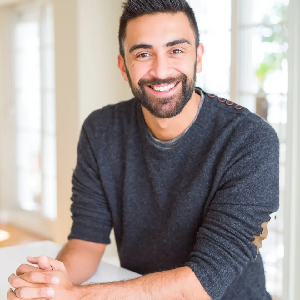 A man with dark hair and a beard, wearing a dark gray sweater, sits at a table and smiles at the camera in a bright room.