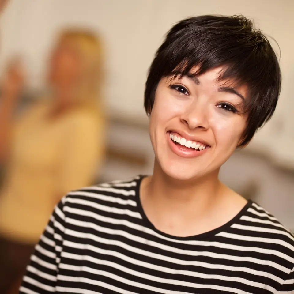 Young woman with short dark hair and striped shirt smiles at the camera, with another person blurred in the background indoors.