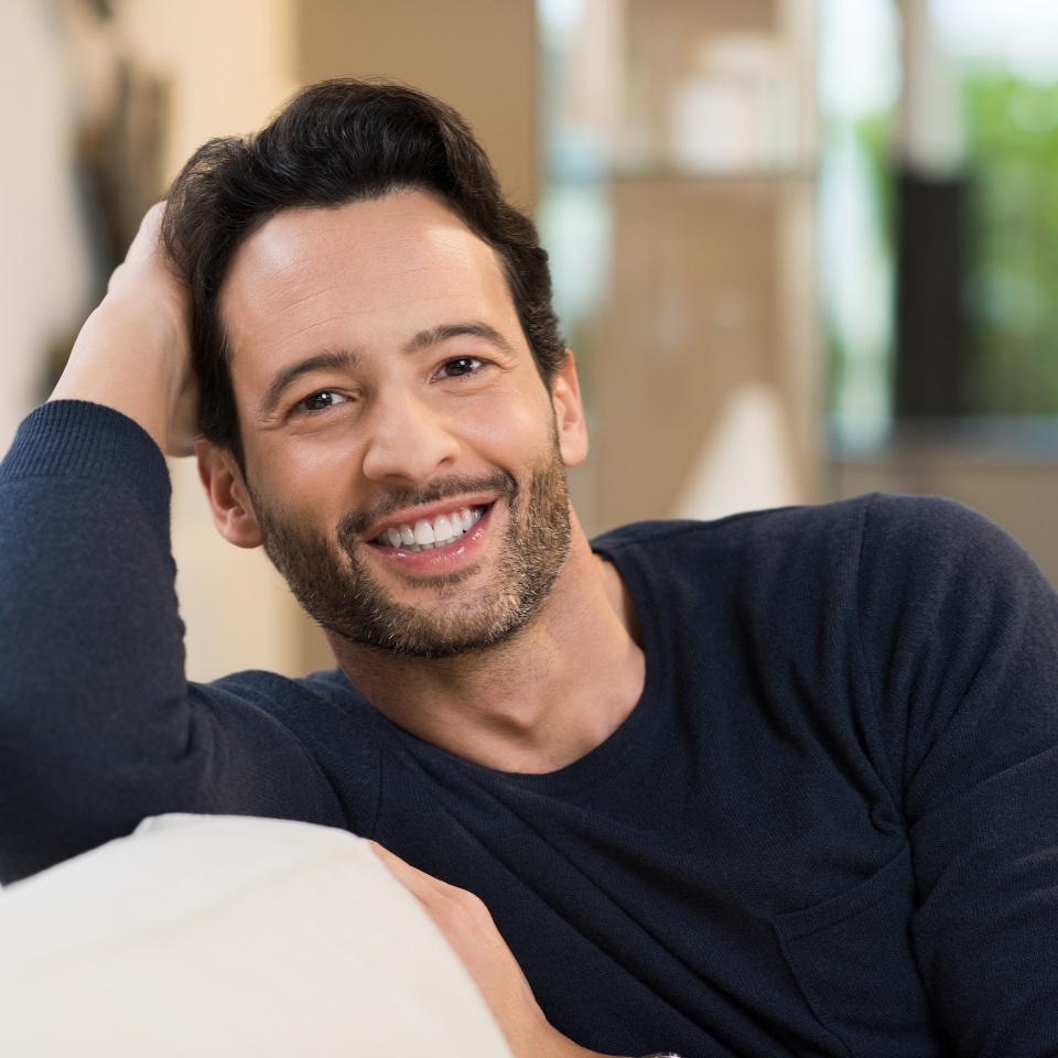 Portrait of a smiling happy young man resting while sitting on sofa. Handsome guy looking at camera. Young latin man in casual relaxing at home. Man relaxing on sofa with arm behind the head.