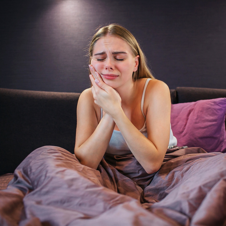 Young woman suffers from toothache. She holds both hands on cheek together and keeps eyes closed. It is painful. Model sits on bed. Her legs are covered with blanket.
