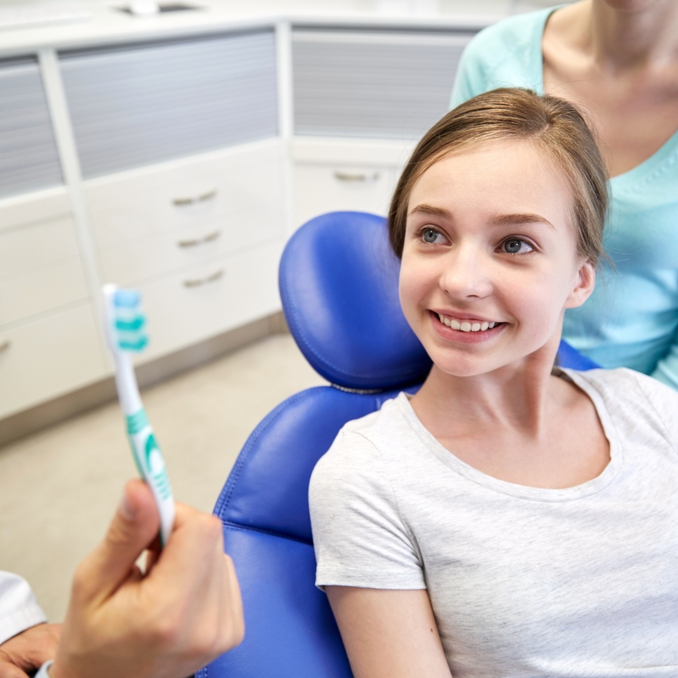people, medicine, stomatology and health care concept - happy male dentist showing toothbrush to patient girl and her mother at dental clinic office