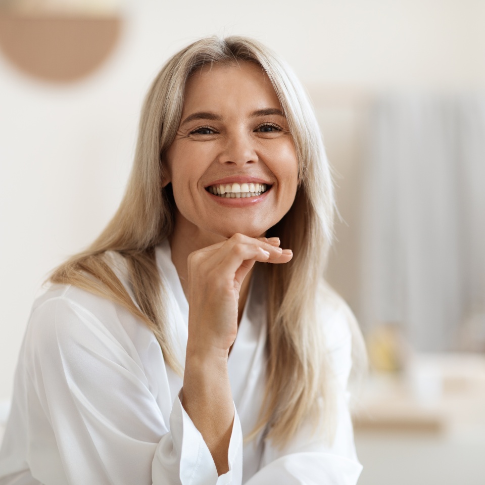 Happy attractive blonde woman in white silky bathrobe posing at white bathroom, smiling at camera, closeup portrait, panorama with copy space. Face and skin care at home concept