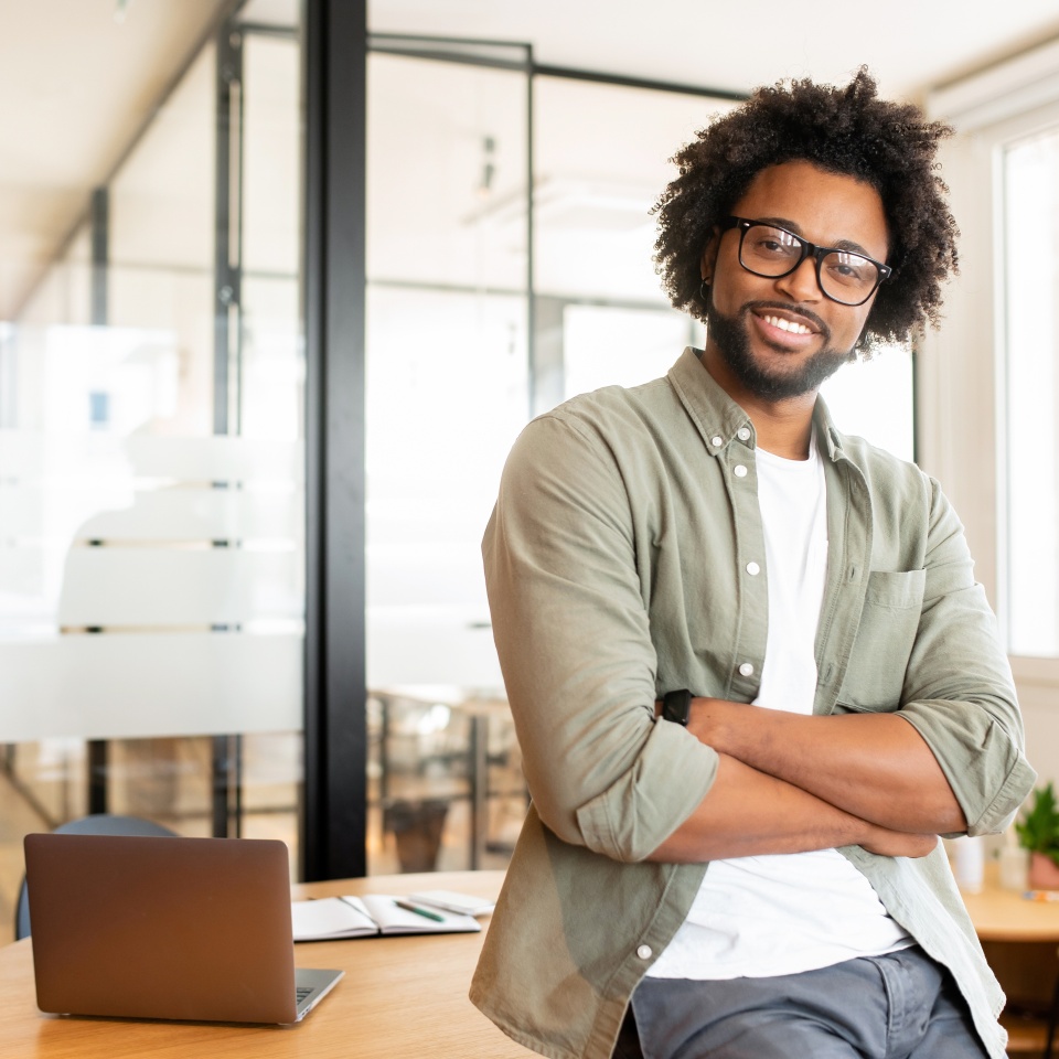 Ambitious highly-skilled handsome curly african-american male employee in glasses and casual wear leaned on office desk and looking at the camera with smile, businessman stands with arms crossed