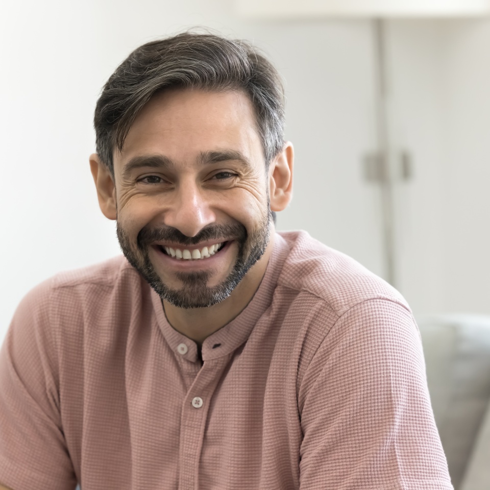 Handsome middle-aged man with warm smile, wear light pink shirt posing for camera, head shot portrait view. Portuguese male looks relaxed, contented, resting alone seated on cozy sofa in living room