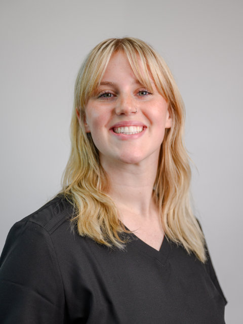 A woman with blonde hair and bangs, wearing a black V-neck shirt, smiles at the camera against a plain light background.