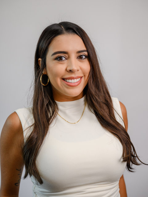 A woman with long brown hair wearing a white sleeveless top, gold hoop earrings, and a gold necklace, smiling at the camera against a plain light background.