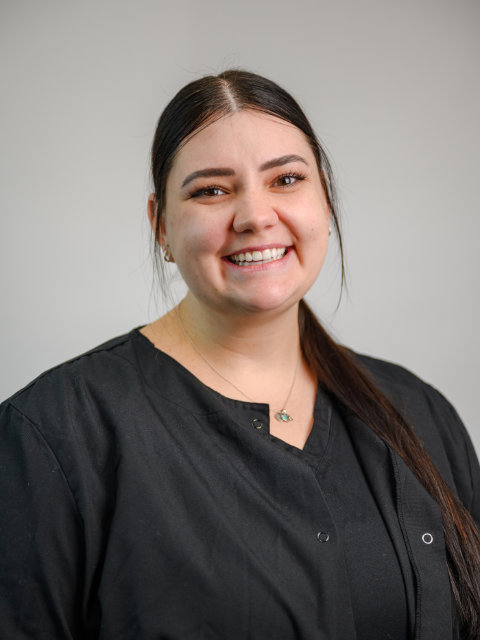 A woman with long dark hair, wearing a black shirt, smiles while standing in front of a plain light gray background.