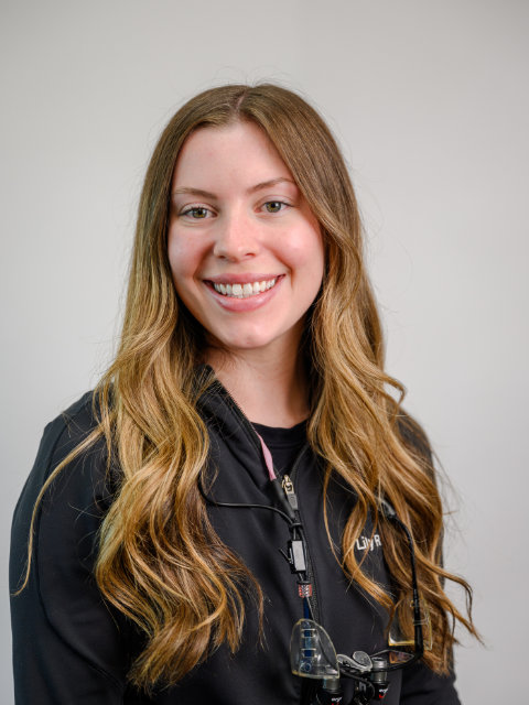 A young woman with long, wavy light brown hair smiles at the camera, wearing a black jacket and dental loupes around her neck against a plain background.
