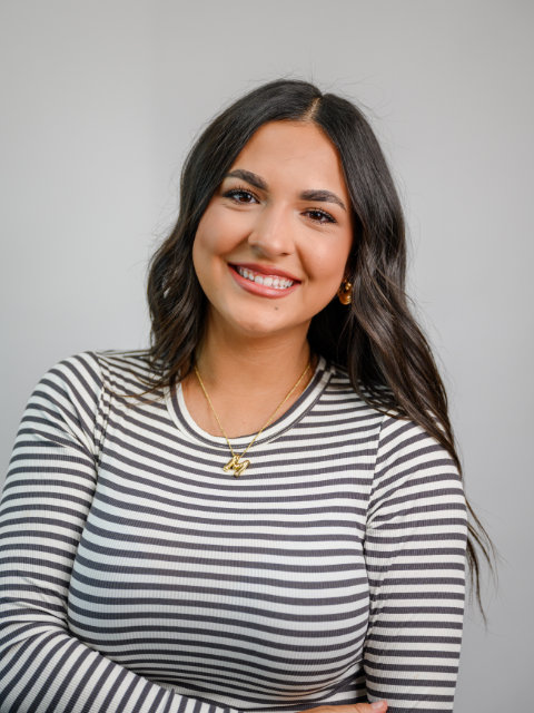 A woman with long dark hair smiles, wearing a black and white striped shirt and gold jewelry, standing against a plain light background.
