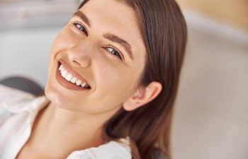 A young woman with straight brown hair smiles while looking up at the camera, showing her teeth. She is wearing a white shirt.