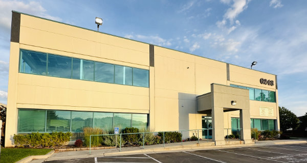 A beige two-story office building with large windows, an entrance canopy, and a mostly empty parking lot in front, photographed in daylight.