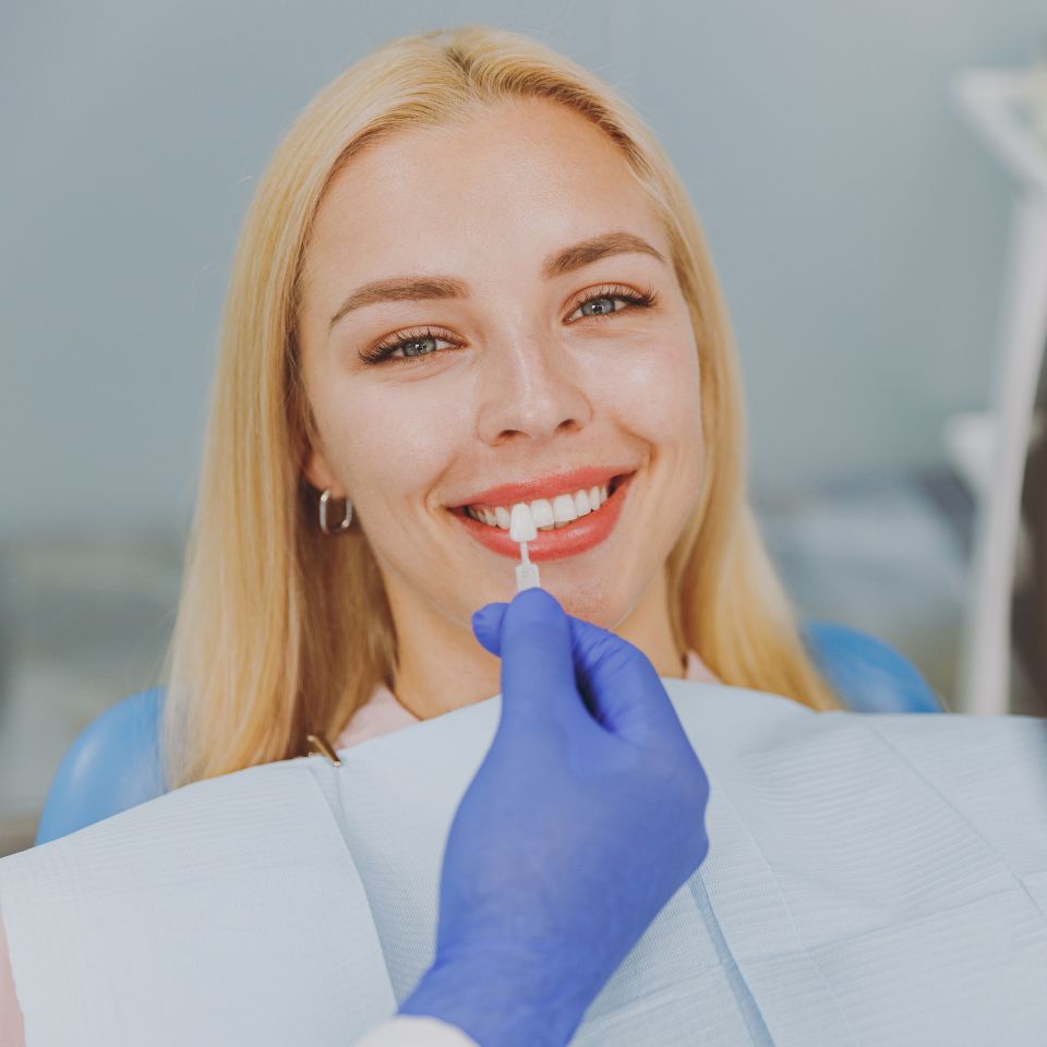 A woman sitting in a dental chair smiles while a gloved hand holds a tooth shade guide to her teeth.
